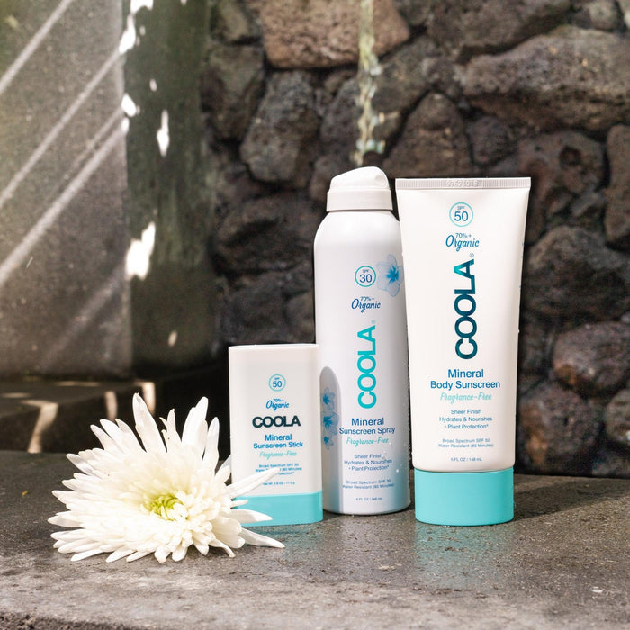 COOLA Mineral Stick SPF 50 displayed alongside sunscreen spray and lotion on a stone surface next to a white flower, promoting organic skincare and sun protection.