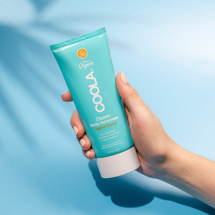 Hand holding a turquoise tube of Classic Body Organic Sunscreen Lotion SPF 30 - Tropical Coconut | COOLA against a light blue background, showcasing skincare and body care product.