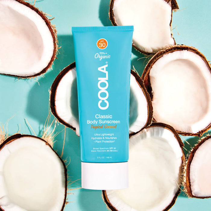 Classic Body Organic Sunscreen Lotion SPF 30 - Tropical Coconut | COOLA tube surrounded by coconut halves on a light blue background, highlighting natural skincare and body care elements.