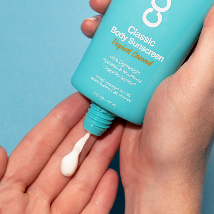 Hand holding a turquoise bottle of Classic Body Organic Sunscreen Lotion SPF 30 - Tropical Coconut | COOLA, dispensing white sunscreen lotion onto the other hand against a blue background.